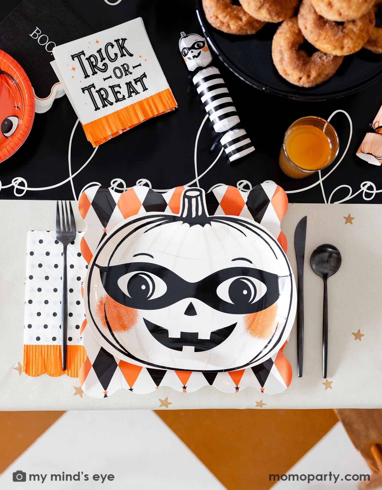 A festive vintage Halloween themed party table setting featuring multiple vintage Halloween themed party supplies from Momopartys including the orange and black harlequin plate, a white vintage Halloween pumpkin shaped plate, the polka dot orange fringe dinner napkin, the trick or treat small napkin, pumpkin party crackers on the boo paper table runner. With drinks, treats, black cutlery set, this fun tablescape is perfect for a kid's friendly Halloween celebration. 