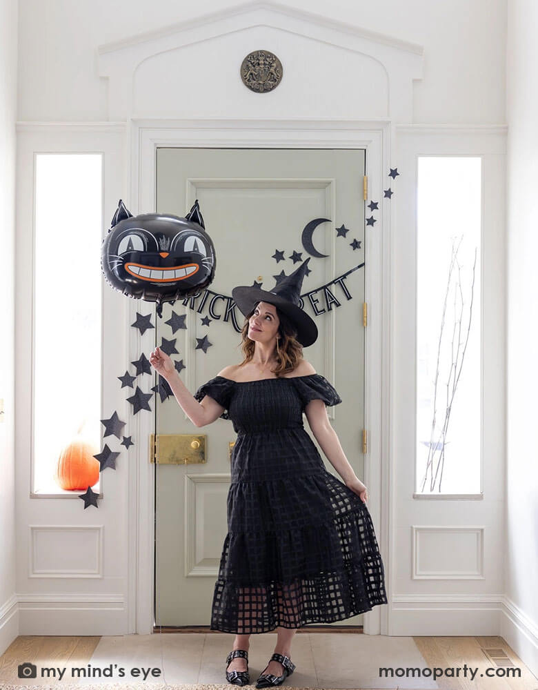 A lady dressed as a Halloween witch holding Momopartys's 24" black cat shaped foil balloon at the doorway decorated with Momopartys's 4ft Trick or Treat felt banner and glittered stars and moon wall paper decorations by My Mind's Eye. Behind the door through the side windows you can see a pumpkin and some spooky branches as the decorations, making this a perfect Halloween home decor idea for this spooky season!