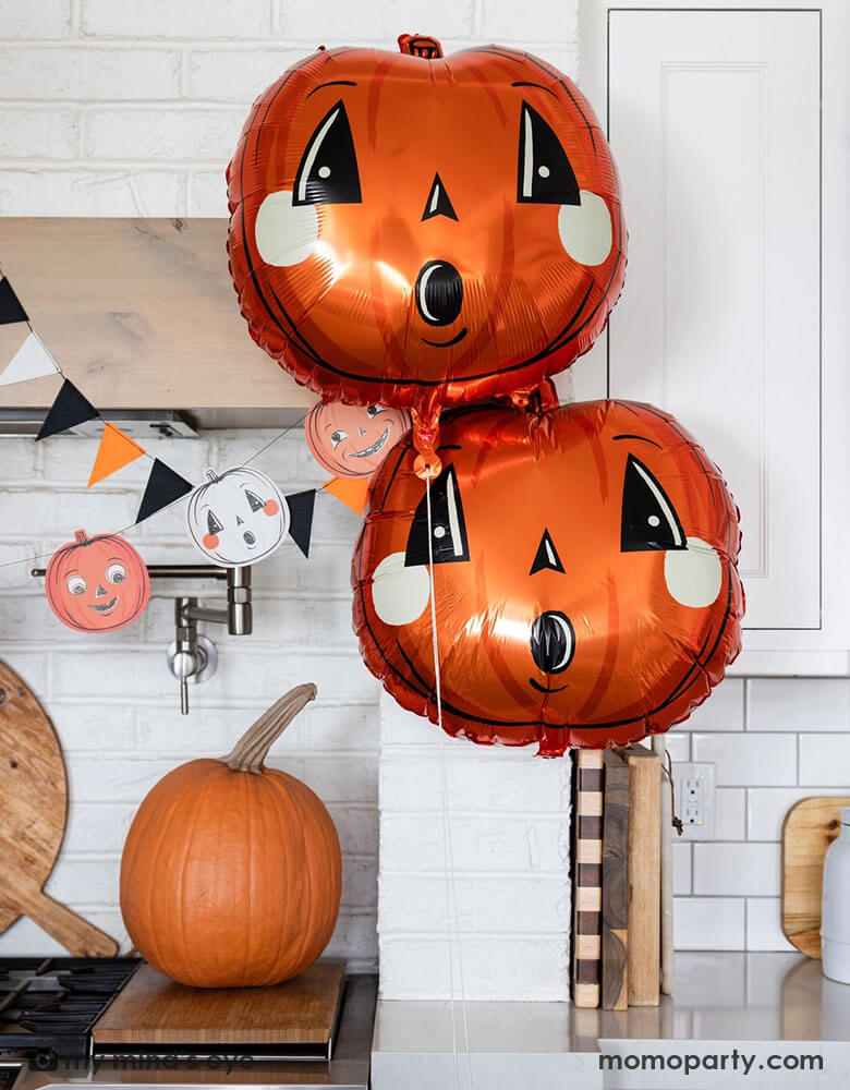 A modern white kitchen decorated for Halloween with Momopartys's vintage Halloween party supplies by My Mind's Eye including pumpkin shaped foil balloons, black, white and orange pennant flags and and paper banner. With a large pumpkin next to the stove, this makes a festive Home decoration to celebrate the spooky season.