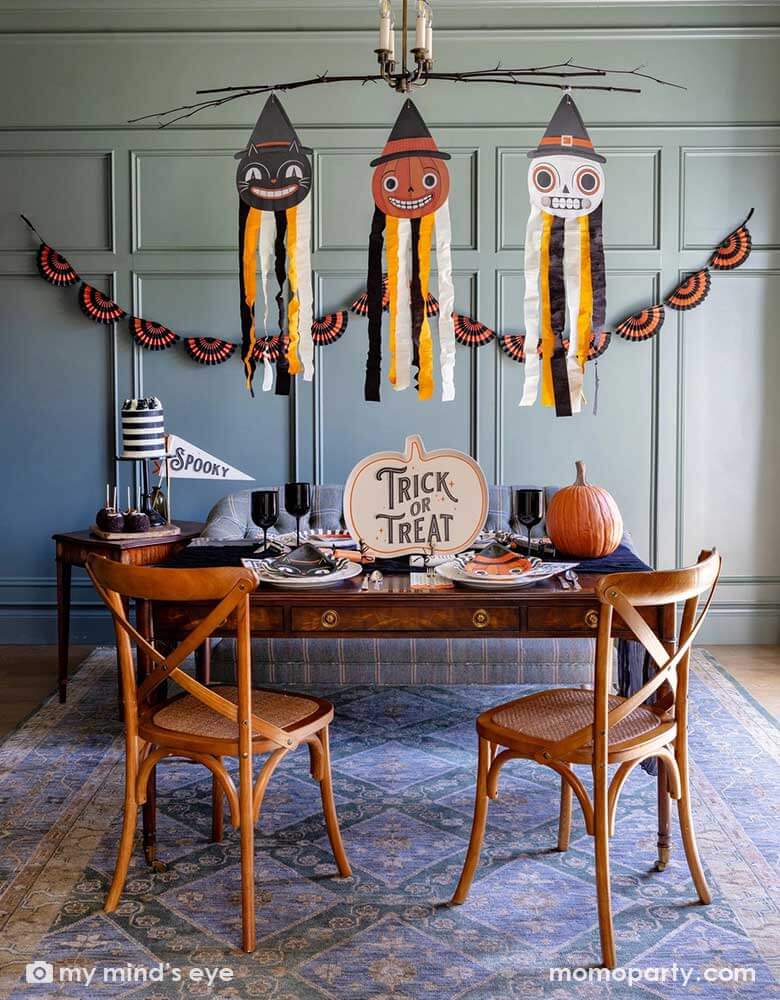 A Halloween party set up featuring vintage Halloween party supplies and decorations from Momopartys including the giant vintage Halloween character hanging decoration streamers and the black and orange bunting banner on the wall as the backdrop. On the table, there's a huge pumpkin shaped bamboo serving plate that says trick or treat on it. With vintage Halloween themed plates, napkins and glasses, along with a black striped cake sitting next to the spooky party pennant - a fun Halloween gathering at home.