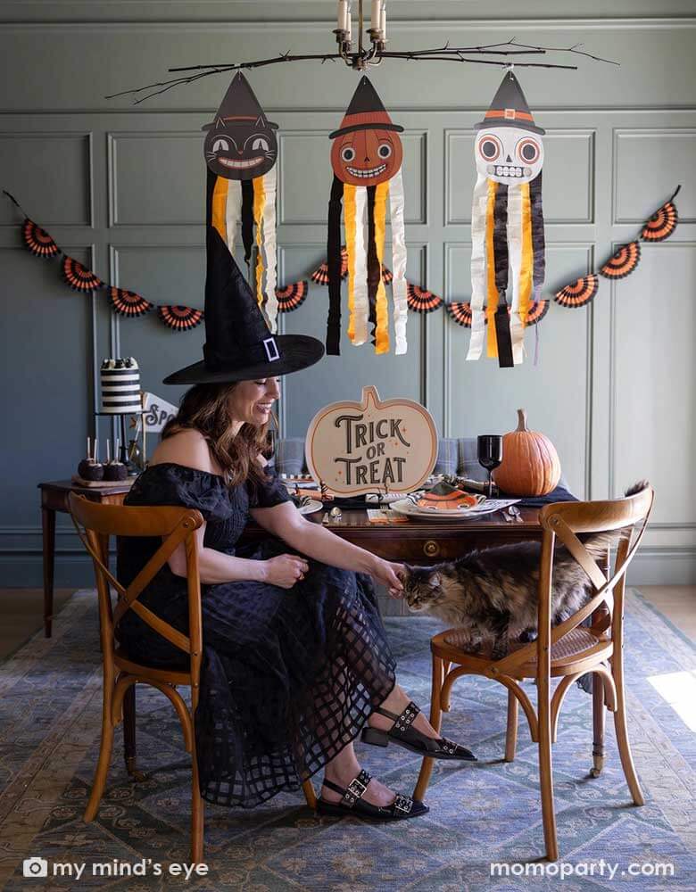 A lady dressed as witch playing with her cat sitting next to her in Halloween party set up featuring vintage Halloween party supplies and decorations from Momopartys including the giant vintage Halloween character hanging decoration streamers and the black and orange bunting banner on the wall as the backdrop. On the table, there's a huge pumpkin shaped bamboo serving plate that says trick or treat on it. With vintage Halloween themed plates, napkins and glasses.