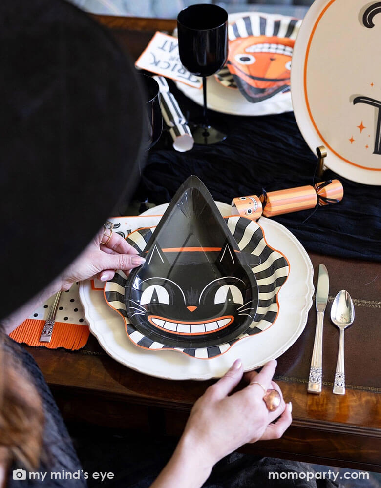A Halloween party table setting featuring vintage inspired Halloween party supplies from Momopartys. The hostess is placing the vintage Halloween black cat shaped plate onto the black starburst large plates and there is the vintage Halloween trick or treat small napkin next to the plates. On the table there are some vintage Halloween character party crackers in the design of a skeleton, a black cat and a pumpkin. Making this a perfect inspo for a festive and spooky Halloween celebration!