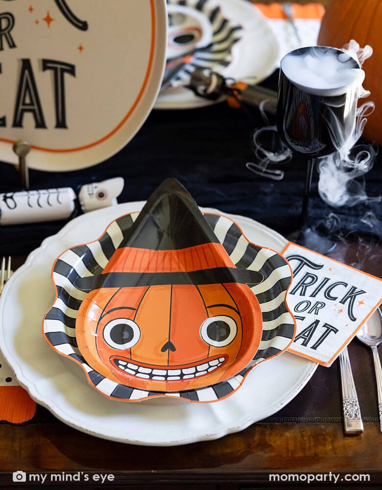 A vintage Halloween party table setting featuring vintage inspired Halloween party supplies from Momopartys including the vintage Halloween pumpkin shaped plate on the black starburst dinner plates and the vintage Halloween trick or treat napkins in vintage fonts. On the table there are some vintage Halloween party crackers in the design of a skeleton, a black cat and a pumpkin. There's also a black wine glass with dry ice as if there's magic smoke coming out for this spooky Halloween celebration!