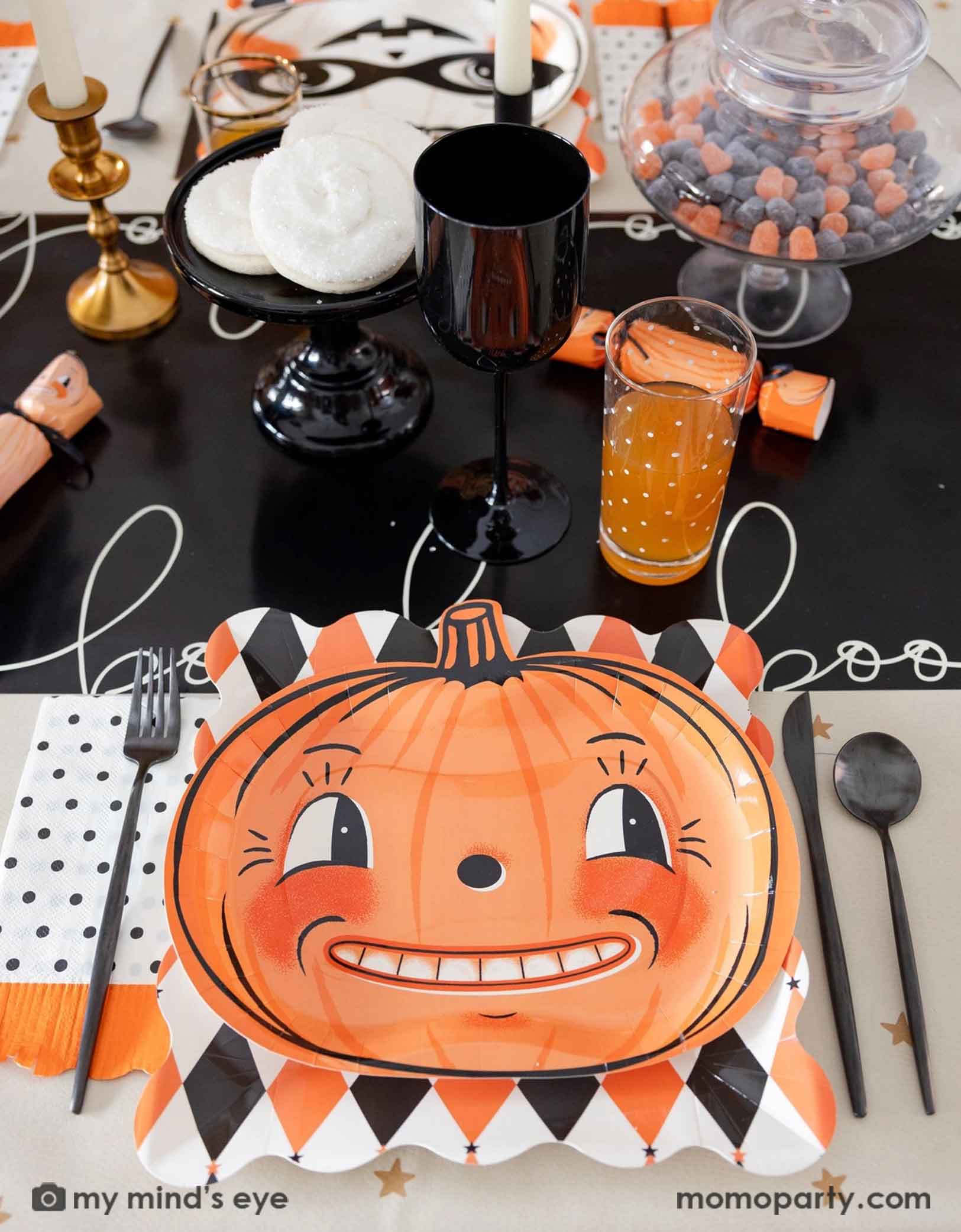 A festive vintage Halloween themed party table setting featuring multiple vintage Halloween themed party supplies from Momopartys including the orange and black harlequin plates, the vintage Halloween pumpkin shaped plates, the polka dot orange fringe dinner napkins, pumpkin party crackers on the boo paper table runner. With drinks, treats, candies and black cutlery set, this fun tablescape is perfect for a kid's friendly Halloween celebration. 