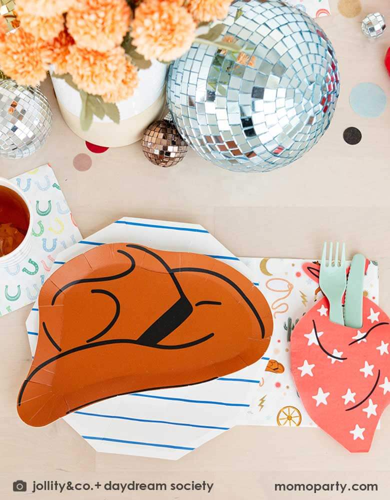A kid's western cowboy themed party table setting feature Momopartys's cowboy themed partyware by Daydream Society including a cowboy shaped plate, a red bandana shaped guest napkin, a Western rodeo themed paper napkin and a blue horseshoe small napkin. Around the tableware there are some fun disco balls around and color coordinated round confetti spread out, with rustic themed flower arrangement as centerpiece, makes this a perfect inspiration for a fun rodeo, cowboy Western themed celebration.