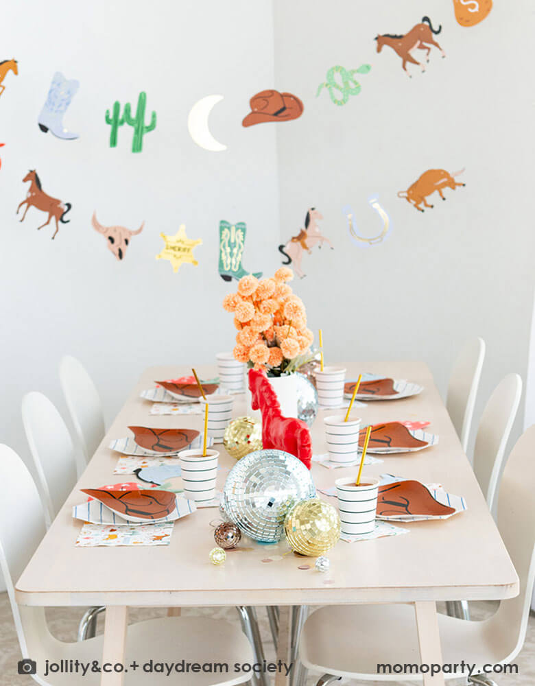 A kid's western themed party set up features Momopartys's cowboy themed party supplies and decorations by Daydream Society including cowboy hat shaped plates, red bandana shaped guest napkins, Western rodeo themed paper napkins and blue horseshoe small napkins. With Western themed party garland featuring pennants including cactus, buffalos, sherif badge, snakes and horses hung above the table, makes this a perfect inspiration for a fun rodeo celebration!