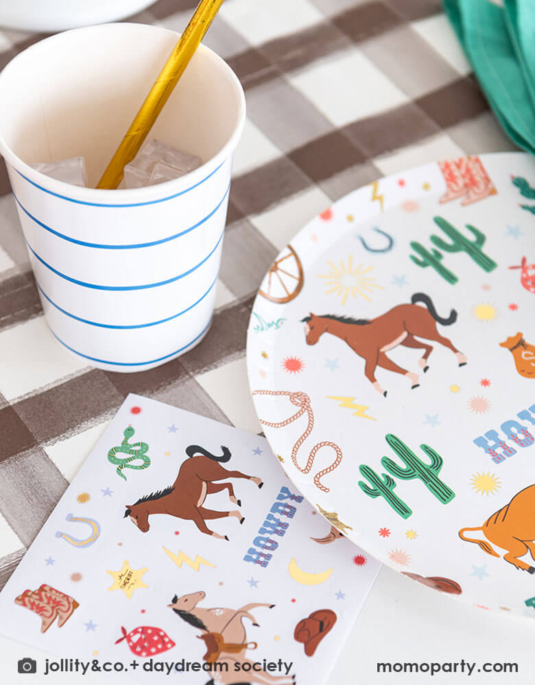 A closeup shot of a western cowboy themed party table tablescape featuring Momopartys's cowboy themed tableware by Jollity Co. and Daydream Society including the round cowboy icon patterned round plate, the small napkin and cactus honeycomb ., all on a brown gingham table runner, making this a modern yet fun tablescape for kid's rodeo, Western or cowboy themed birthday celebration. A perfect inpso for kid's "First Rodeo" first birthday celebration.  