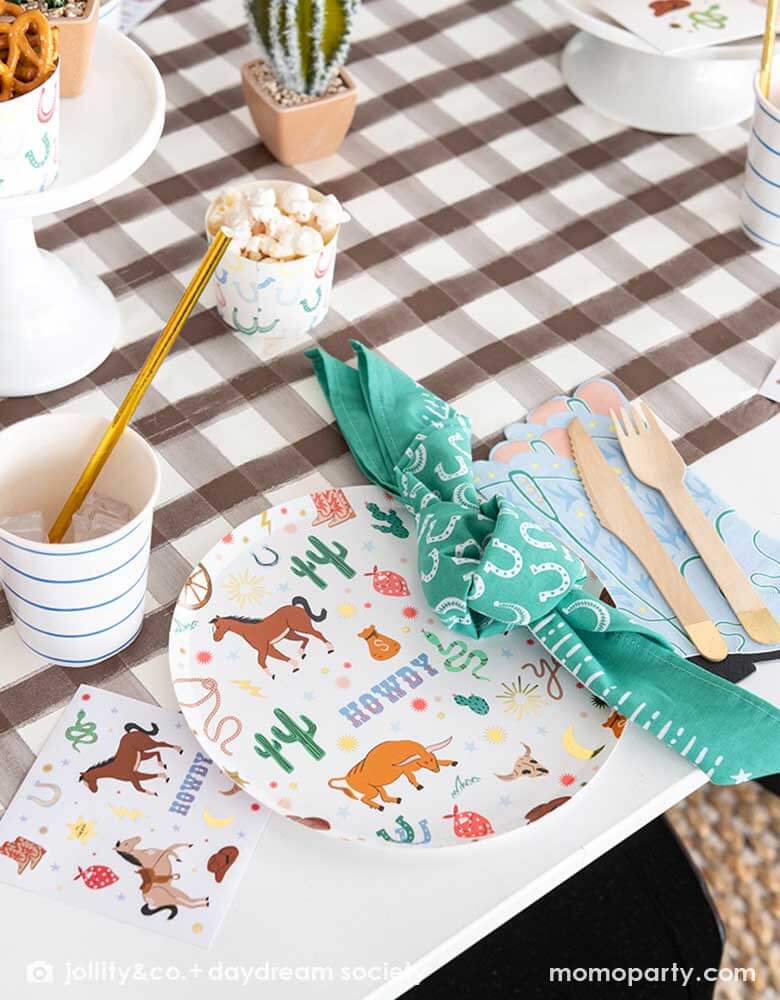 A western cowboy themed party table tablescape featuring Momopartys's cowboy themed tableware by Jollity Co. and Daydream Society including the round cowboy icon patterned round plate, the small napkin, the cowboy boot shaped napkin, horseshoe napkin all on a brown gingham table runner, making this a modern yet fun tablescape for kid's rodeo, Western or cowboy themed birthday celebration. A perfect inpso for kid's "First Rodeo" first birthday celebration.  