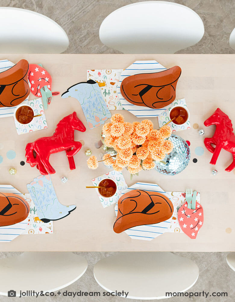 A kid's western themed party table features Momopartys's cowboy themed partyware by Daydream Society including cowboy hat shaped plates, red bandana shaped guest napkins, Western rodeo themed paper napkins and blue horseshoe small napkins. Around the tableware there are some fun disco balls around and color coordinated round confetti spread out, with rustic themed flower arrangement and red horse decorations as the centerpiece, makes this a perfect inspiration for a fun rodeo, cowboy themed celebration.