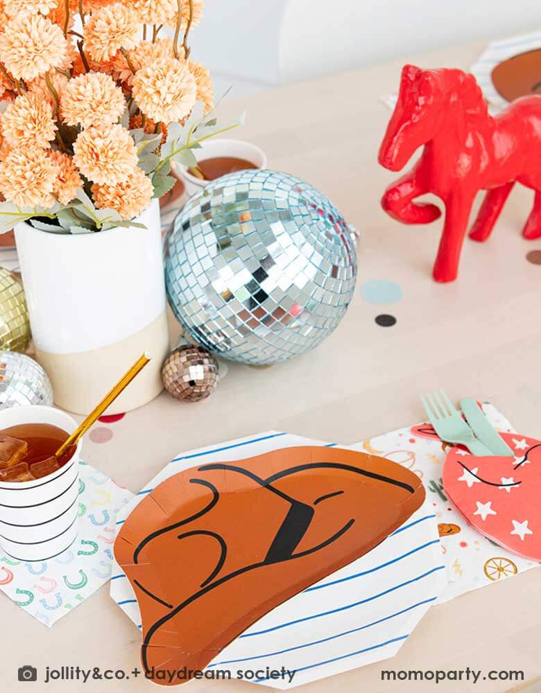 A kid's western themed party table features Momopartys's cowboy themed partyware by Daydream Society including a cowboy shaped plate, a red bandana shaped guest napkin, a Western rodeo themed paper napkin and a blue horseshoe small napkin. Around the tableware there are some fun disco balls around and color coordinated round confetti spread out, with rustic themed flower arrangement and a red horse decoration as the centerpiece, makes this a perfect inspiration for a fun rodeo, cowboy themed celebration.