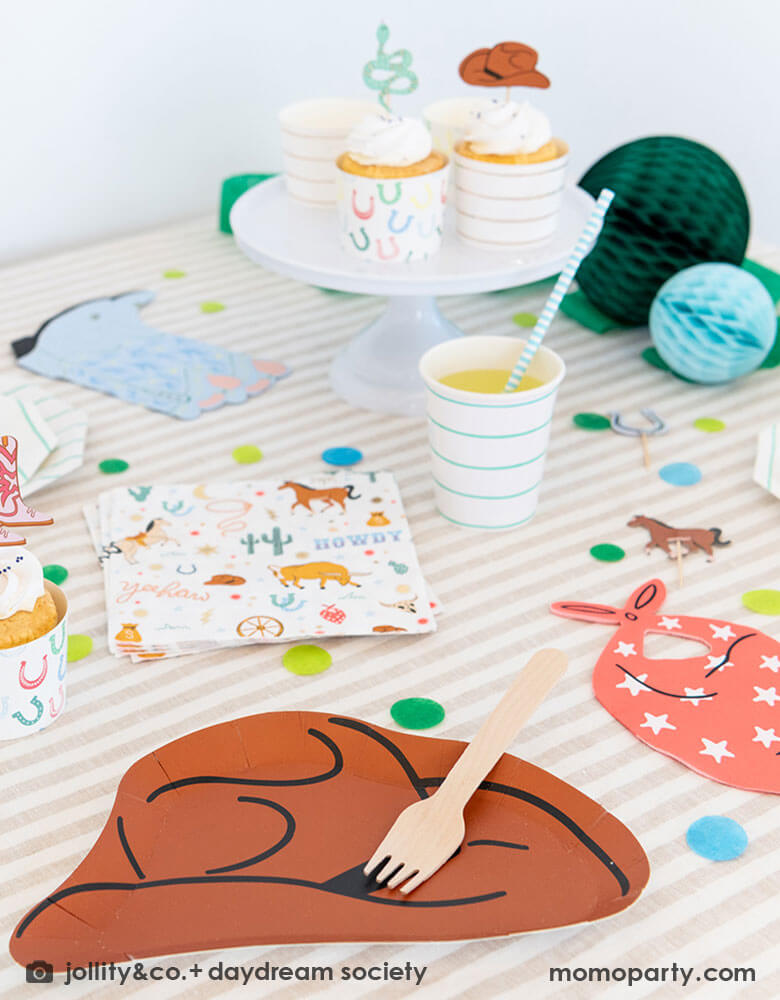 A kid's western themed party table features Momopartys's cowboy themed partyware by Daydream Society including a cowboy shaped plate, a red bandana shaped guest napkin, a Western rodeo themed paper napkin and a blue cowboy boots shaped napkin. On the table there are some cupcakes decorated with cowboy themed toppers, with blue tone honeycomb decorations in back and color coordinated round confetti spread out, this makes a perfect inspiration for a fun rodeo, cowboy themed celebration.