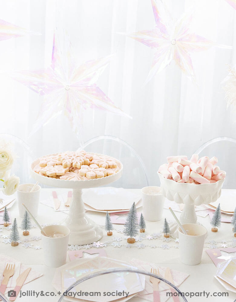 A whimsical winter wonderland themed holiday party table set up featuring Momopartys's frosted party collection including plates, napkins and party cups in snowflake design with iridescent accents. On the table there are some light pink meringue and other desserts. With iridescent star party decorations hung above the table and mini silver bottlebrush Christmas trees on the table, this makes a great inspiration for a girl's dreamy winter themed celebration.