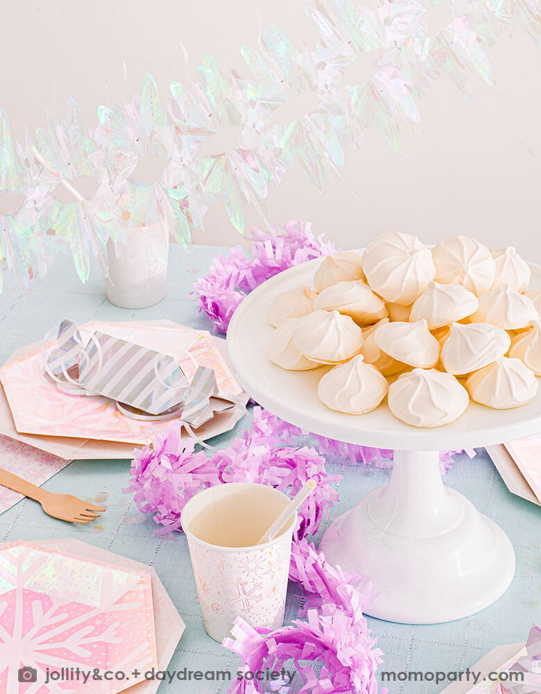 A whimsical winter wonderland or Frozen themed party table set up featuring Momopartys's frosted party collection including plates, napkins and party cups in snowflake design with iridescent accents. On the table there are some light pink meringue desserts and silver candy shaped treat boxes. With iridescent party garland above the table and lilac pink streamers on the table, this makes a great inspiration for a girl's dreamy winter themed celebration.