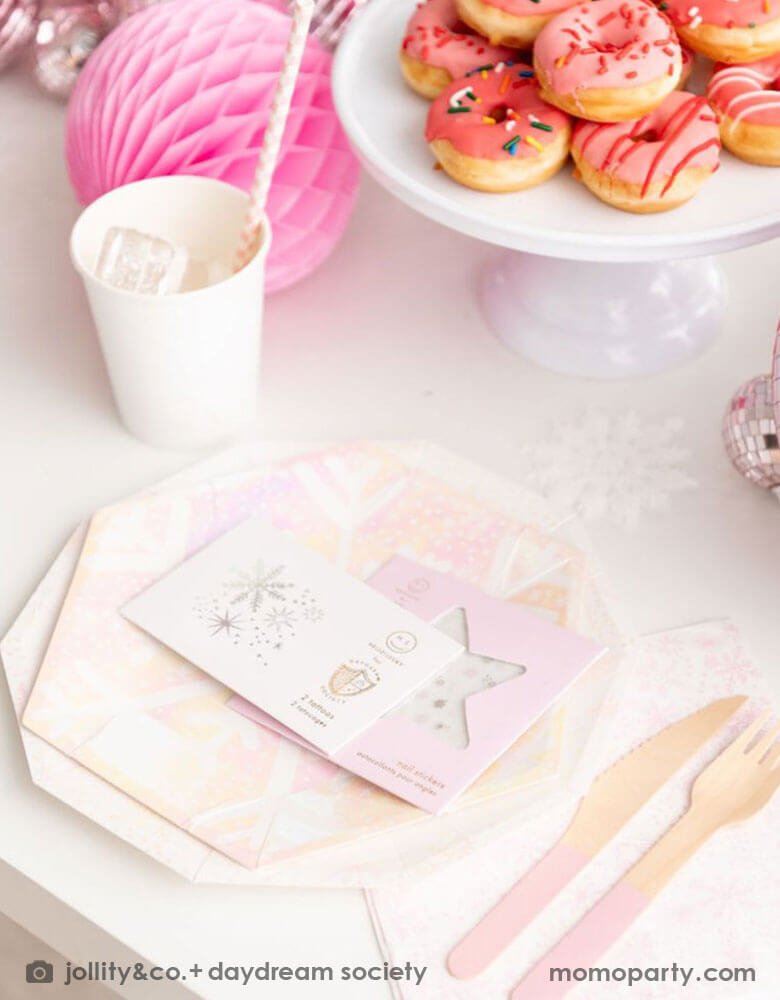 A close up of a whimsical winter wonderland or Frozen themed party table place set up featuring Momopartys's frosted party collection including plates, napkins, party cups, temporary tattoos and nail stickers in snowflake design with iridescent accents. On the table there are some mini donuts in pink color and some pink honeycombs and disco balls as decorations - makes this a great inspiration for a girl's dreamy winter themed celebration.