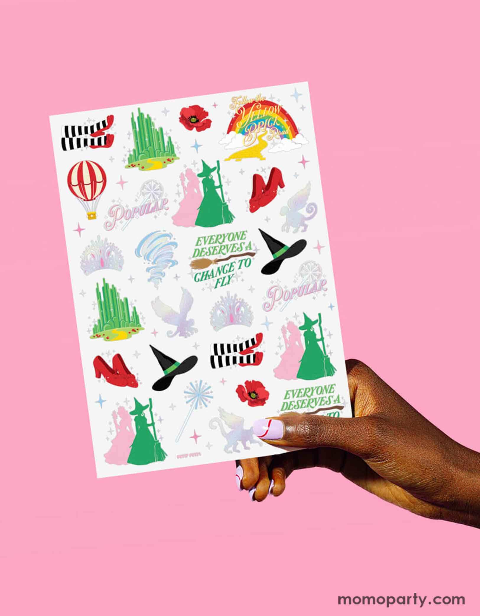 A hand holding Momopartys's Emerald City Wicked Witch Temporary Tattoo sheet by Xo, Fetti. Featuring  green and pink witches, witch hats, broomsticks, Emerald city, wands, tiaras and magical icons inspired by the Wizard of Oz. On the table around the tattoo sheets there are some rainbow shaped plates and party cups with pink juice, making it perfect for a Wicked themed birthday party celebration.