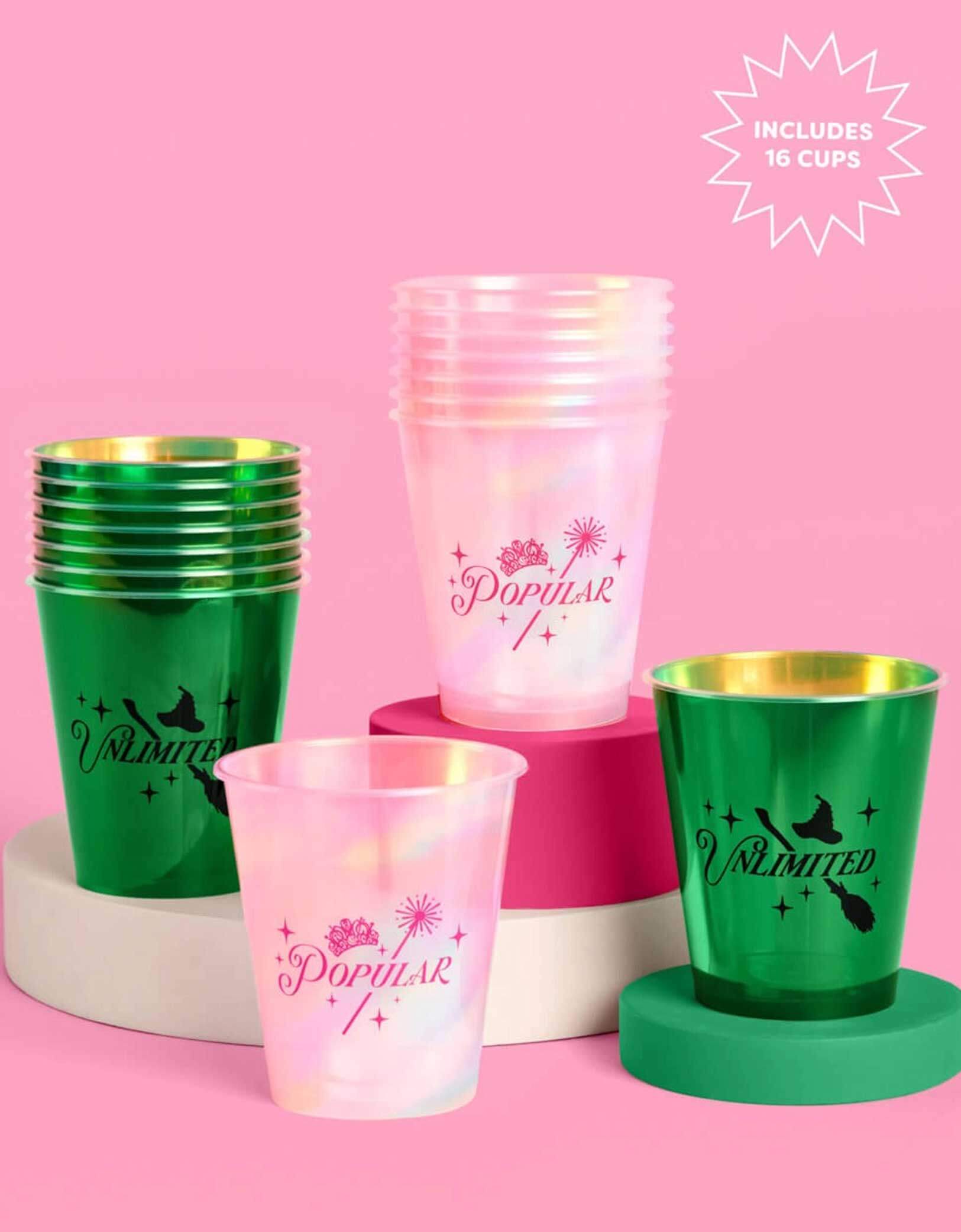 A set of Wicked themed party plastic cups from Momopartys, one set of pink cups with "popular" text on it and the other set of green plastic cups with "unlimited" text on it, on a pink platform with a pink background.