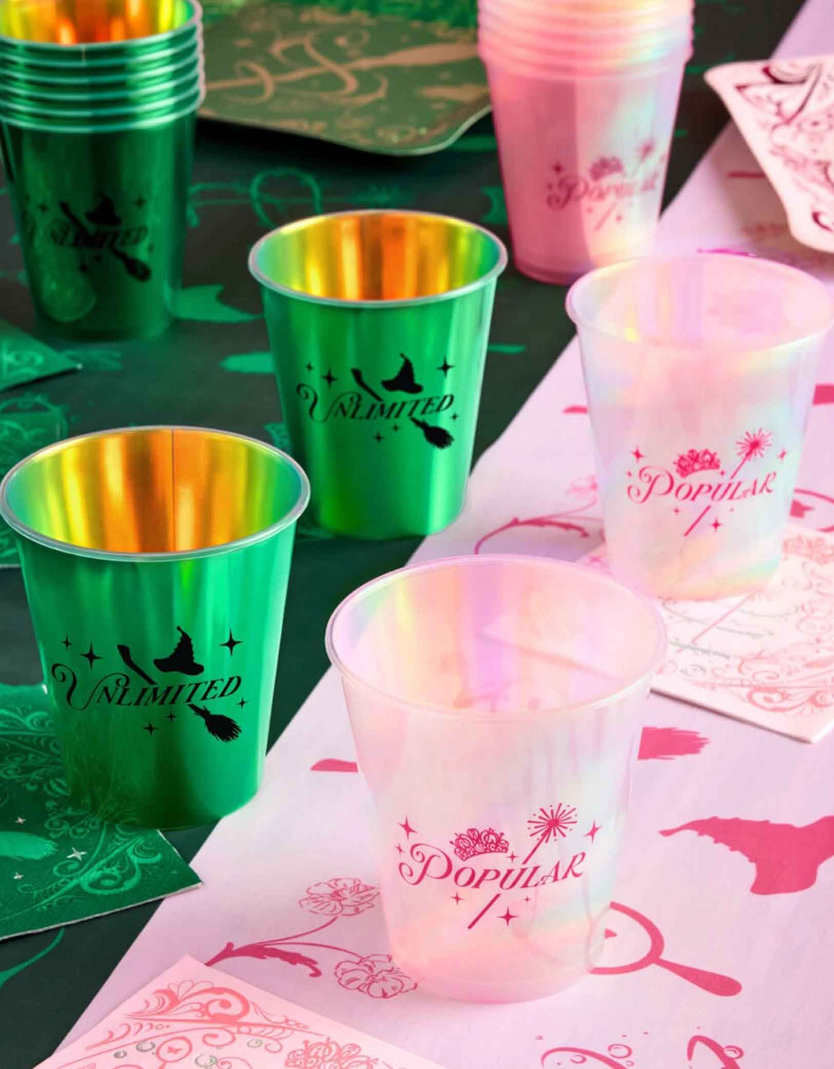 A Wicked themed party table featuring various Wicked themed party supplies from Momopartys including Wicked reusable iridescent plastic cups in pink and green, Wicked two-toned tablecloth, Wicked party dinner plates and napkins in green and pink with wizard themed pattern design on them.