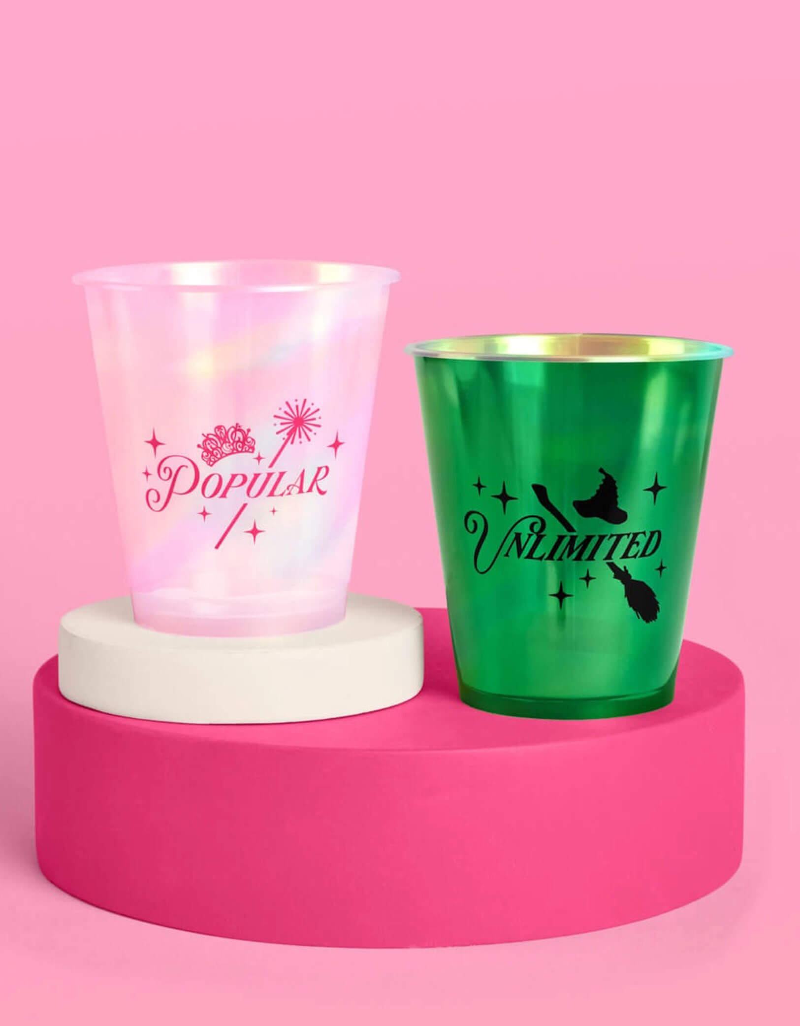 Two Wicked themed party plastic cups from Momopartys, one pink with "popular" text on it and one green with "unlimited" text on it, on a pink platform with a pink background.