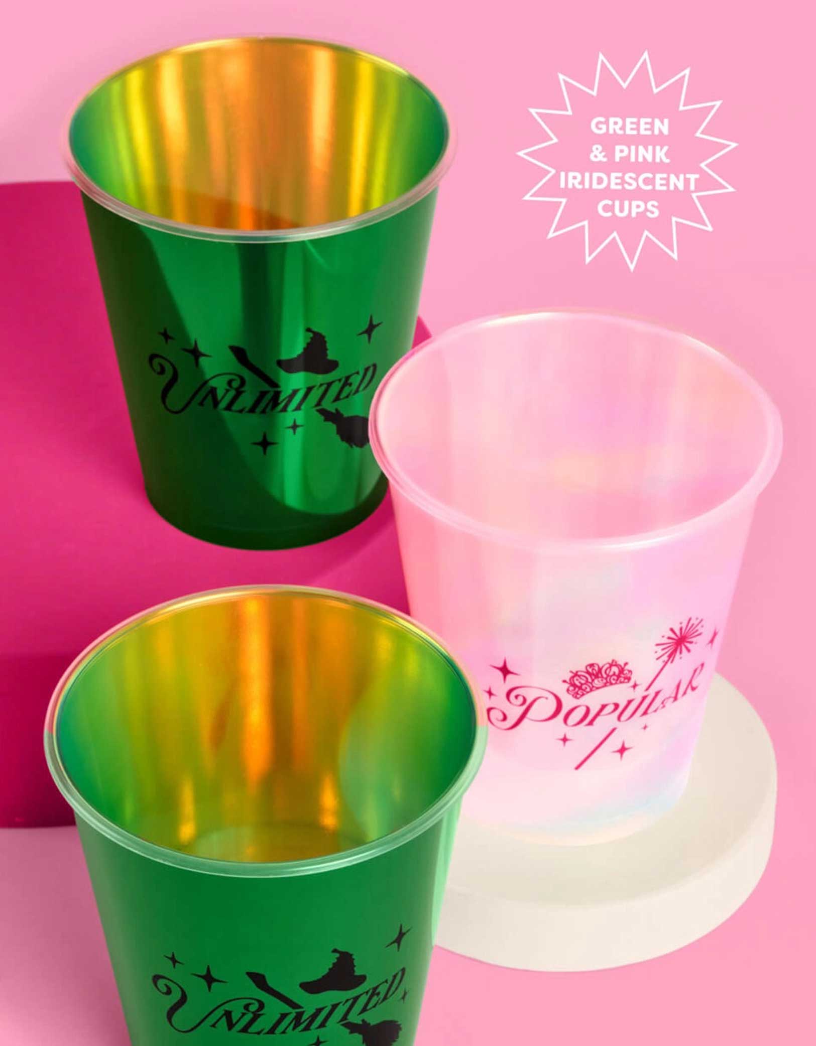 Three Wicked themed party plastic cups from Momopartys, one pink with "popular" text on it and two green cups with "unlimited" text on it, on a pink platform with a pink background.