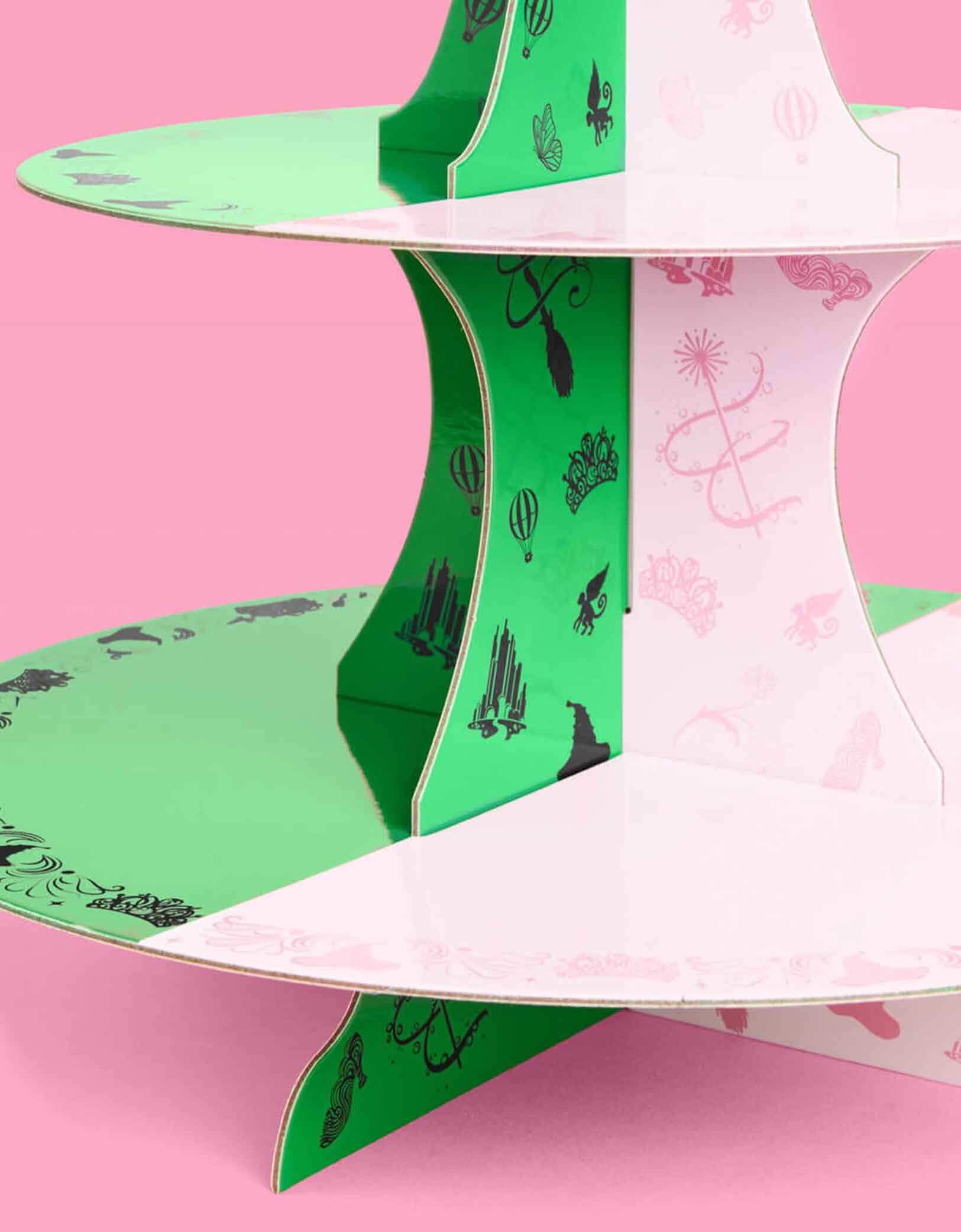 A close up shot of Momopartys's green and pink cake stand with Wicked's Emerald City design at the top on a pink background. Inspired by the enchanting world of the Wicked Witch, this green and pink centerpiece featuring tiaras, wands, hot air balloons, witch hats, and more add a whimsical flair to a Wicked themed celebration. Perfect for showcasing your sweet treats, it transforms your dessert table into a dazzling scene straight from the Emerald City.