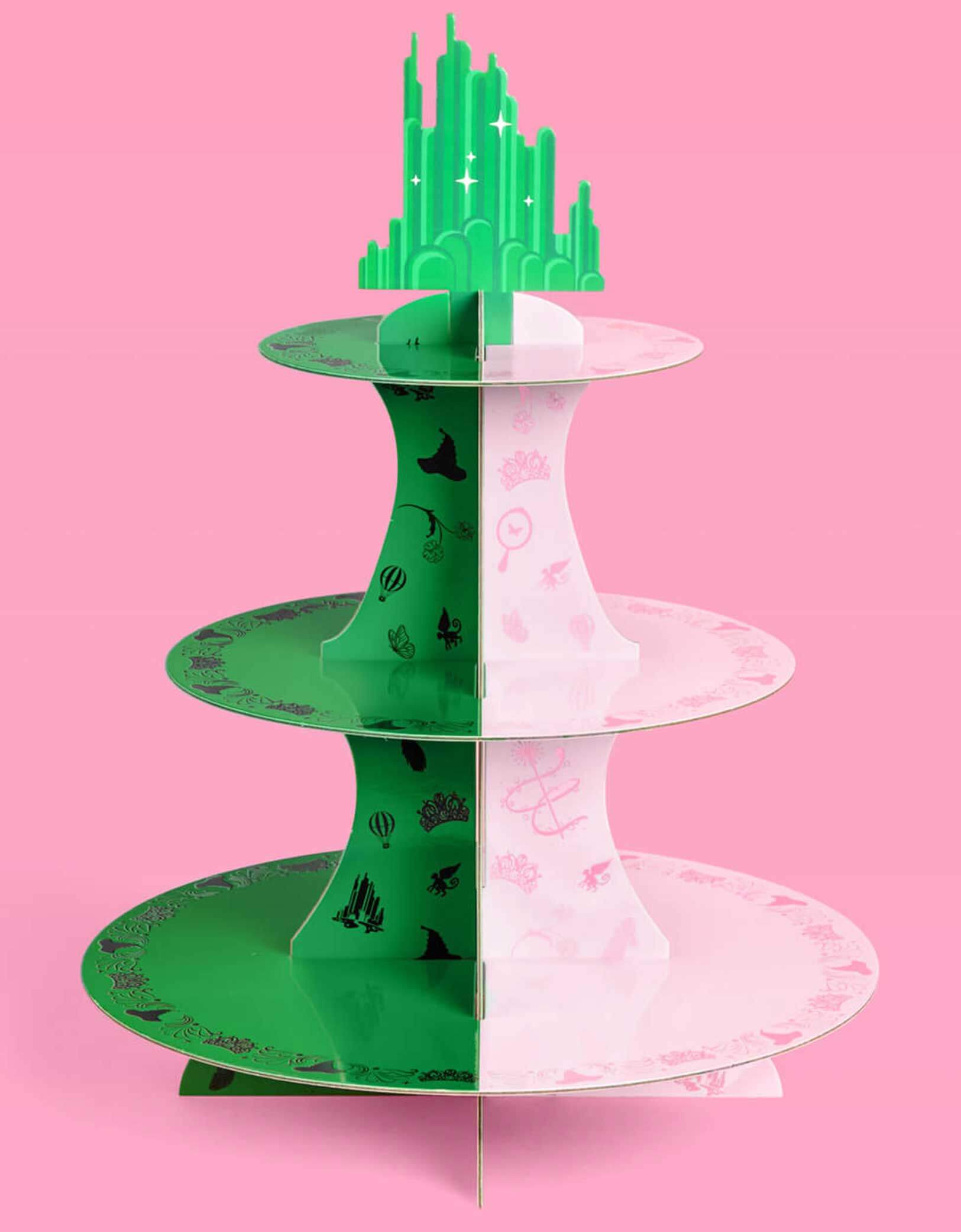 Momo Party's green and pink cake stand with Wicked's Emerald City design at the top on a pink background. Inspired by the enchanting world of the Wicked Witch, this green and pink centerpiece adds a whimsical flair to any celebration. Perfect for showcasing your sweet treats, it transforms your dessert table into a dazzling scene straight from the Emerald City.&nbsp; Topped with a green cityscape with iridescent foil details, let your cupcakes shine and your guests be charmed by this playful, magical displ