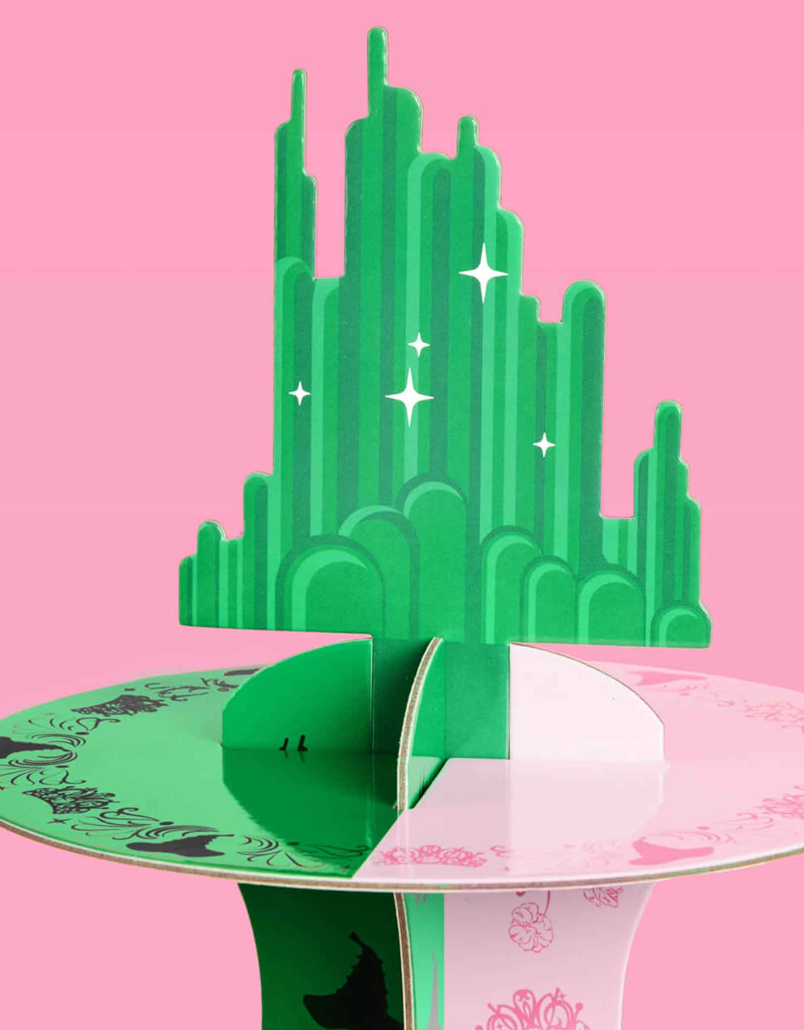 A close up of the Emerald City Topper on Momopartys's green and pink Wicked cake stand on a pink background. Inspired by the enchanting world of the Wicked Witch, this green and pink centerpiece adds a whimsical flair to any celebration. Perfect for showcasing your sweet treats, it transforms your dessert table into a dazzling scene straight from the Emerald City.