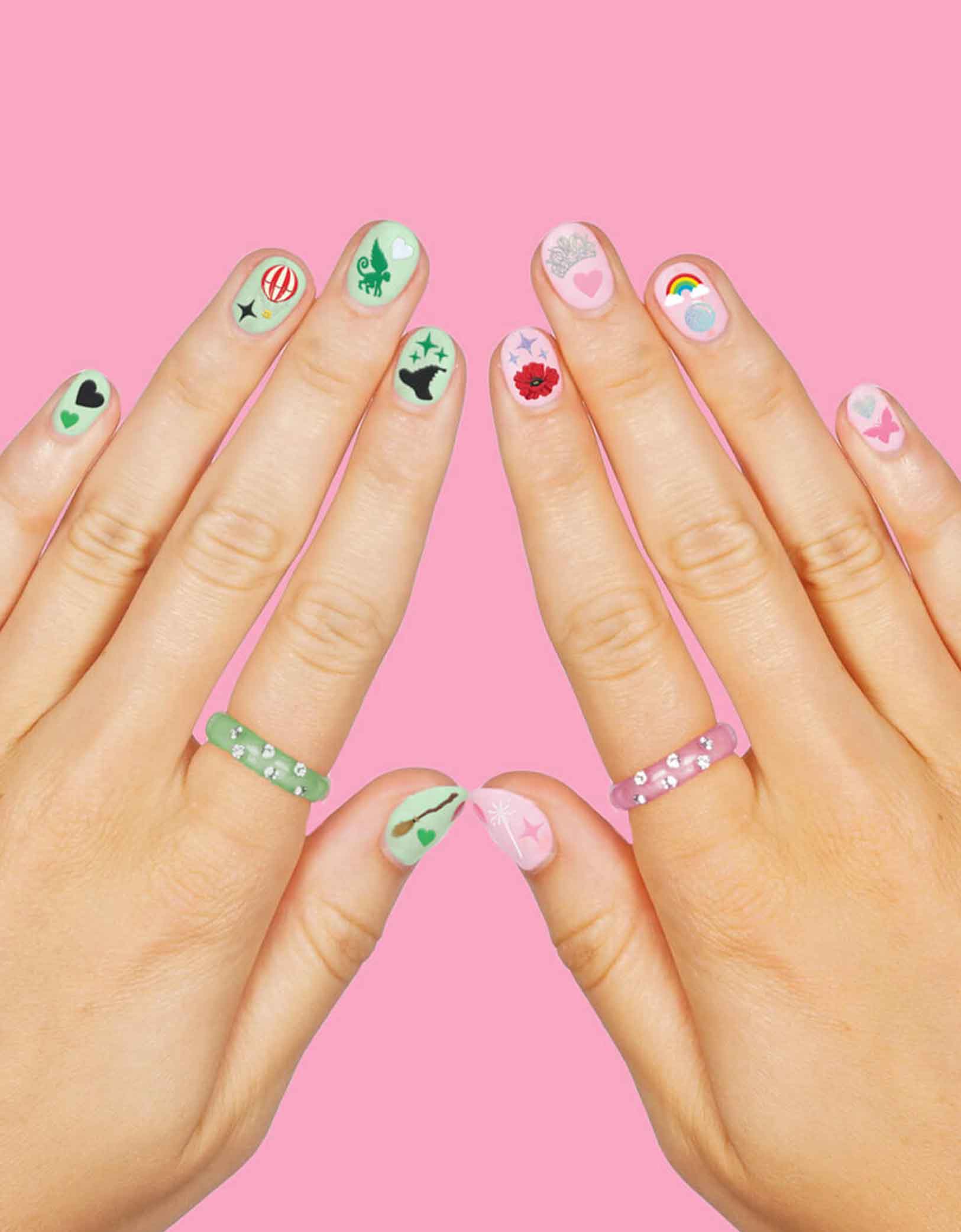 Two hands with with Momopartys's Wicked Emerald City Nail Stickers by Xo, Fetti on a pink background, featuring wicked themed elements including witch hats, bubbles, hot air balloons, butterflies, tiaras, rainbows, sparkles, witch's brooms,  they’re perfect for a fun, themed activity that lets guests express their playful side. Easy to apply and remove, these stickers bring a splash of fantasy to your fingertips, making every celebration a little more spellbinding.