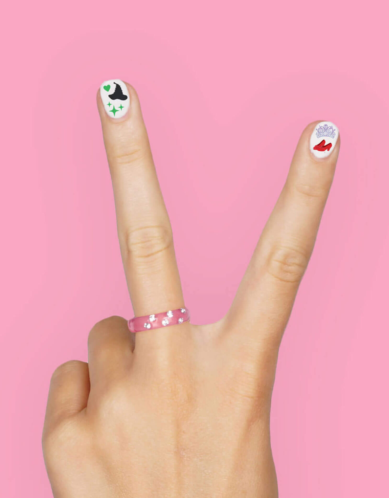 Hand making a peace sign with decorated Wicked themed nail stickers from Momopartys against a pink background.