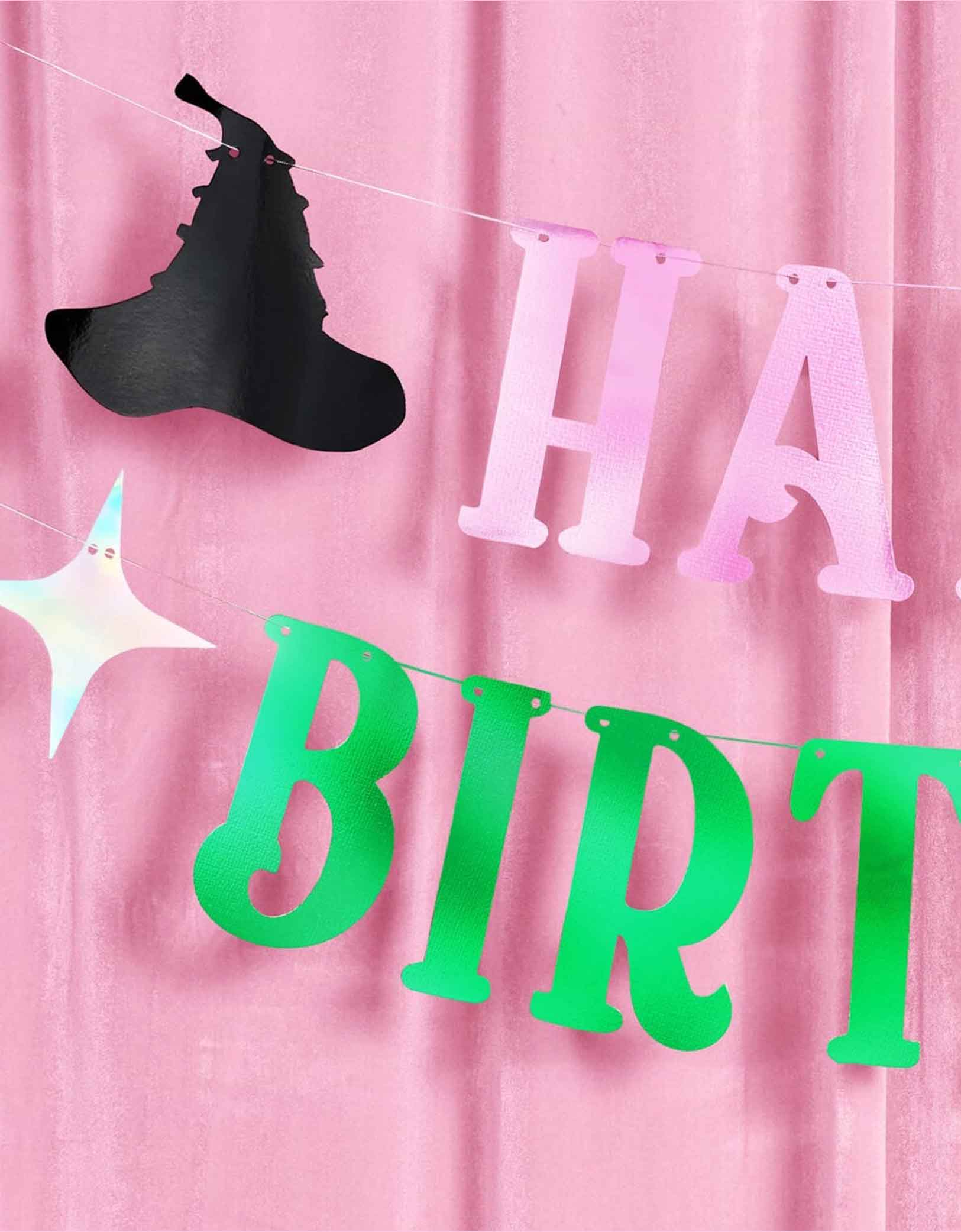 A closeup of Momopartys's 4ft Wicked pink and green Happy Birthday banner with decorative elements including an iridescent tiara, a witch hat and star point pennants on a pink curtain background. 