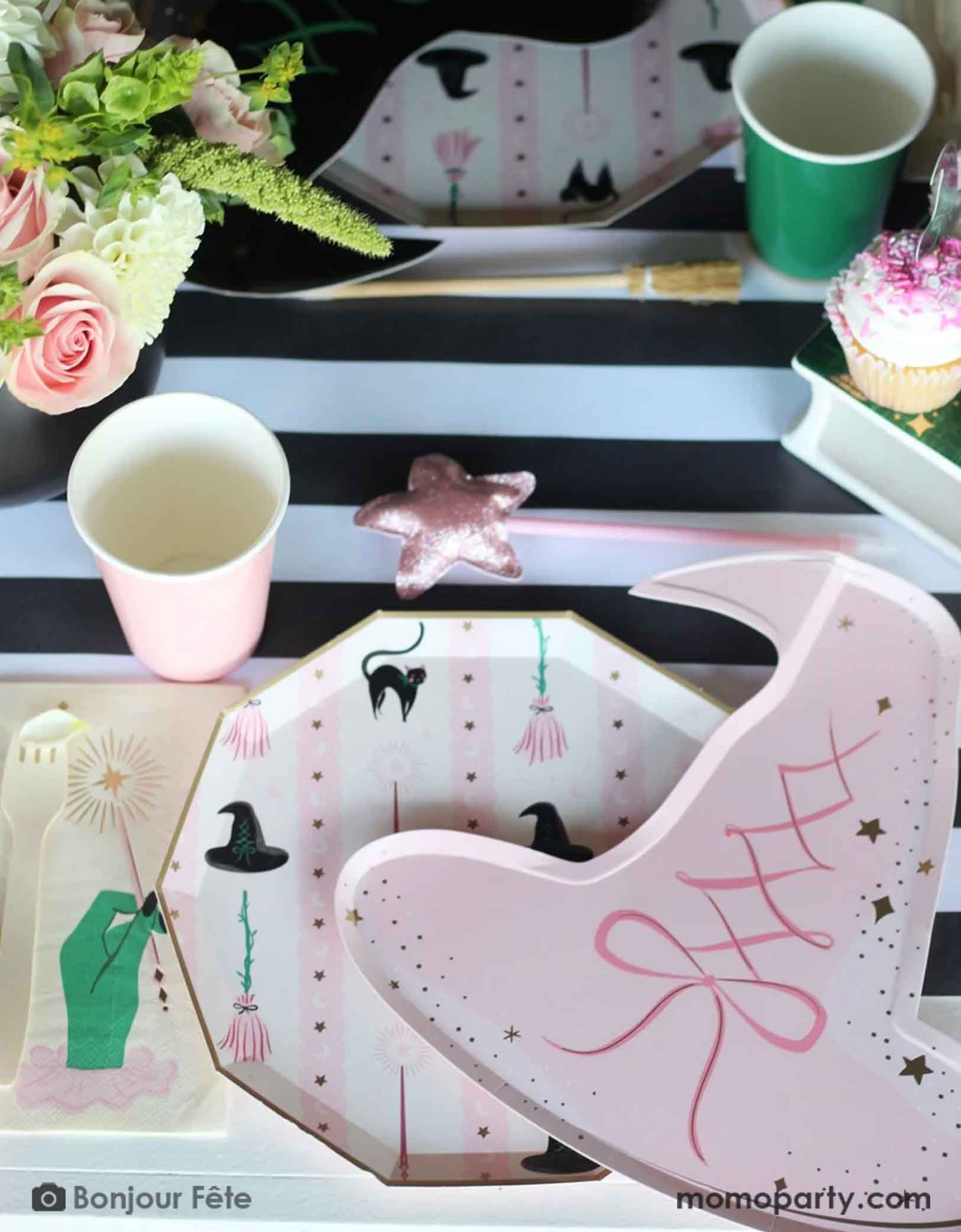 A whimsical Wicked themed party table setting featuring multiple Wicked themed party supplies from Momopartys including the Pink Witch Hat shaped Plate, with Wicked themed side plate and matching napkin, along with Wicked party cups in green and pink on a black striped table runner underneath. With floral arrangement and a pink star wand, it's a perfect party setting for a fun and magical Wicked themed party for both kids and adults.