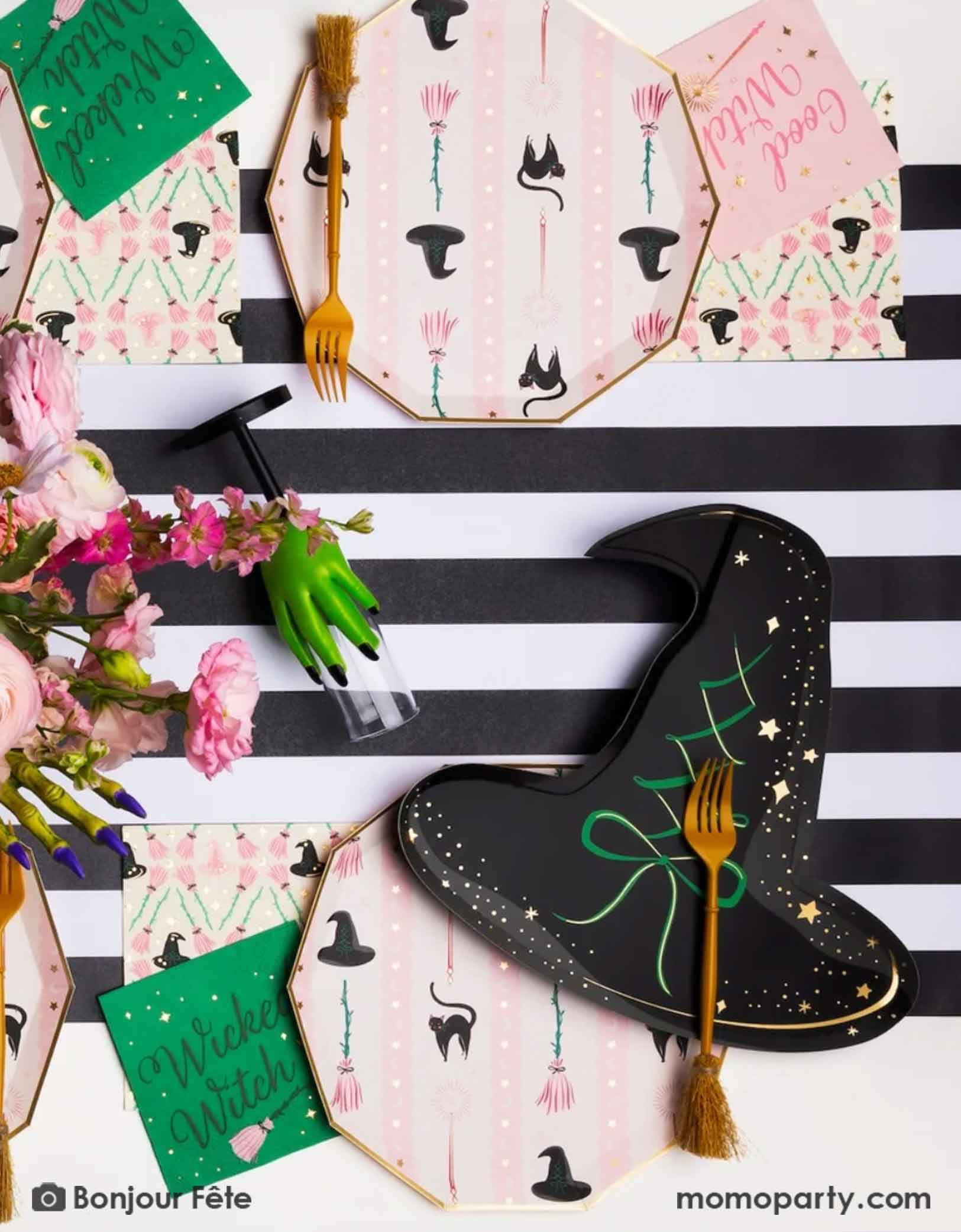 A whimsical Wicked themed party table setting featuring multiple Wicked themed party supplies from Momopartys including the Wicked Witch Hat shaped Plate, the 5" small napkins, the witch's broom shaped utensils, the large pink witch themed dinner plates, along with Wicked themed drinking glasses with a green hand and a black striped table runner underneath confetti spread out on the table and a mini, it's a perfect party setting for a fun and magical Wicked themed party in the Halloween spooky season.