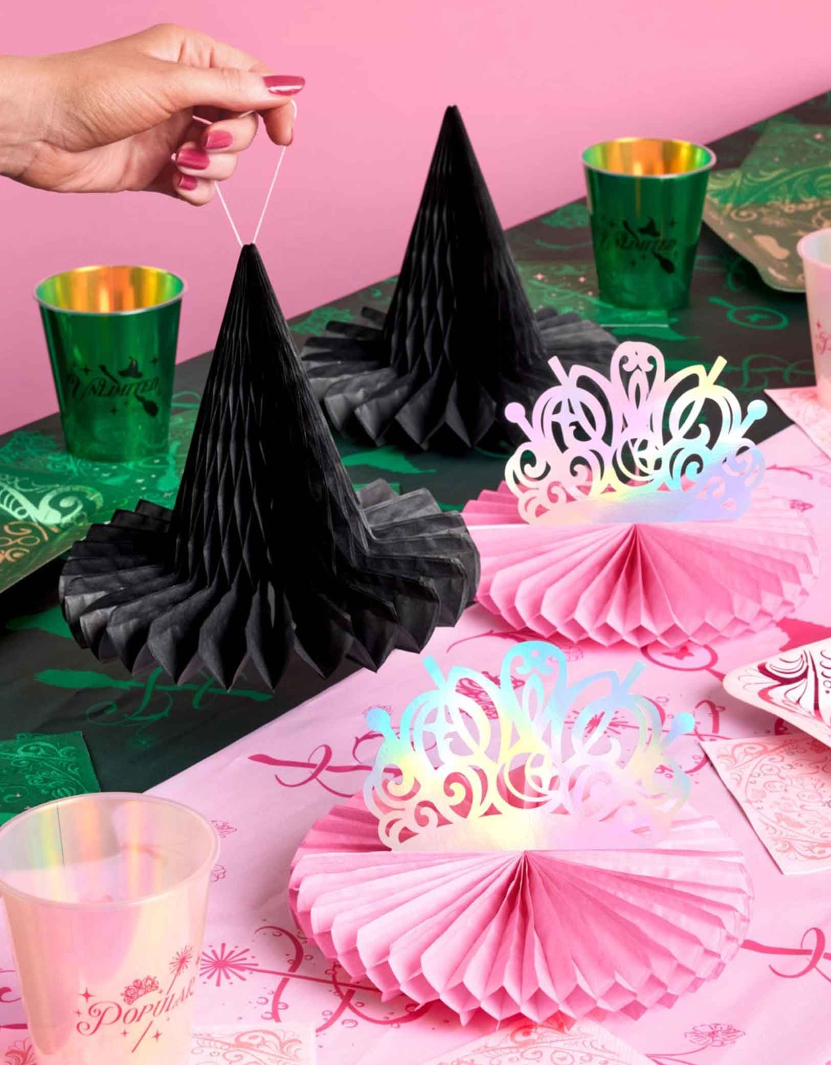 A festive Wicked themed party table featuring various of Wicked themed tablewares and decorations from Momopartys including Wicked Tiara & Witch Hat Honeycomb Set, Wicked party cups in green and green and Wicked party plates in green and pink on a Wicked themed tablecloth.