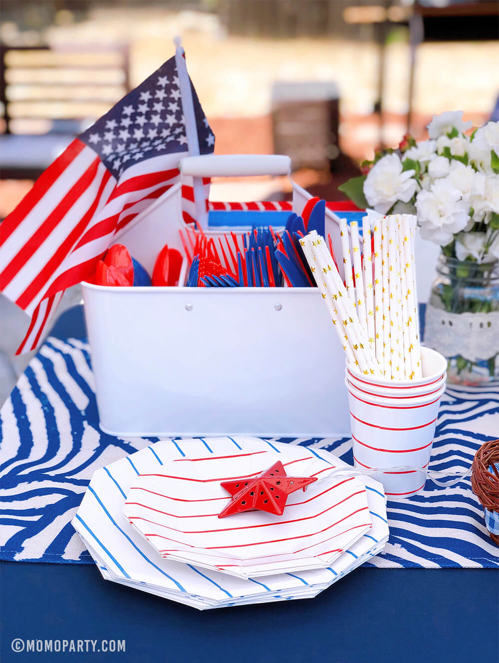 Morden backyard 4th of July Party table set up with Day dream society Blue Striped Large Plates, Red Striped Small Plate, Red Striped Cups, gold start print paper straw, American flag in the cutlery container, flowers on a blue strip table runner.  