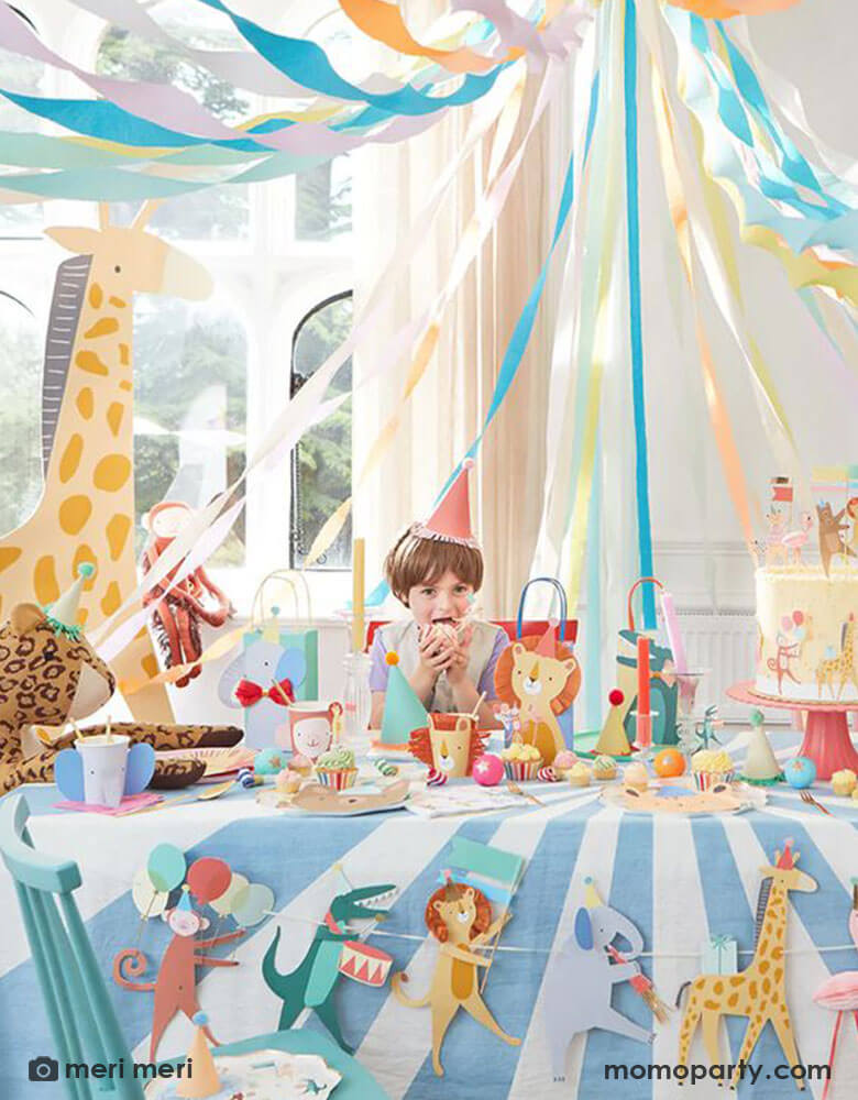 A festive party set up for kid's animal themed party featuring Momopartys's Animal Parade collection by Meri Meri including a jumbo sized animal parade garland, animal shaped party cups featuring an elephant, a monkey, an alligator, and a lion with colorful party bags with animal designs, cupcake toppers and cake toppers, a perfect idea for kid's "Wild One" themed 1st birthday party and "Two Wild" 2nd birthday party.