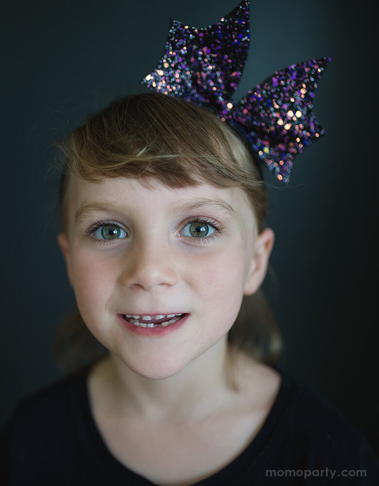 Girl wearing Great Pretenders - Bat Bow Headband. This sparkly bat bow headband in purple color will add an extra Halloween flair to any outfit! Beautiful and affordable Accessory for girl, Accessory for kid sold by party boutique online store momopartys.shop