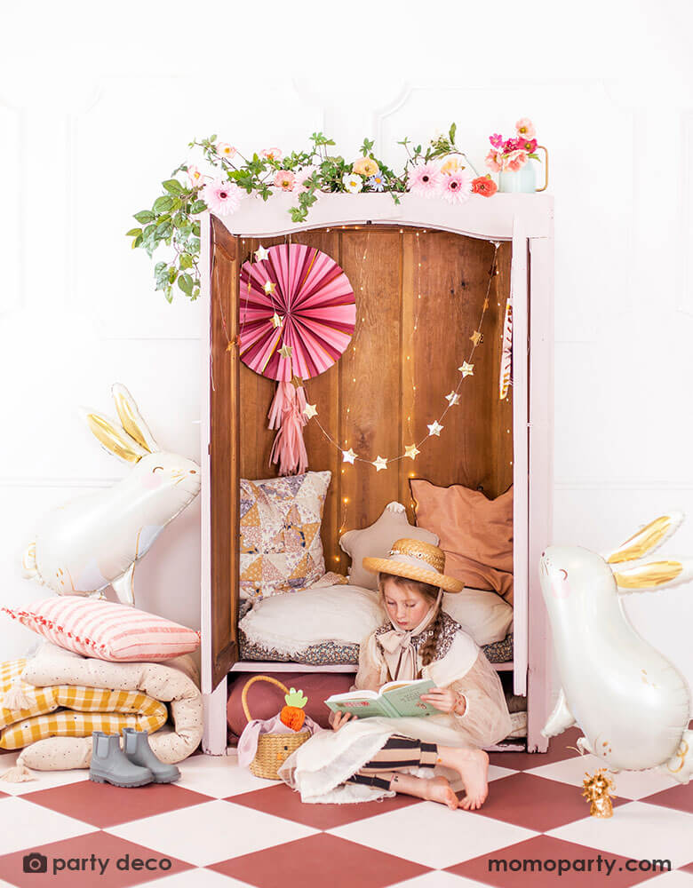 A closet decorated with beautiful spring flowers and a pink party fan with tassels and gold star banner. Next to the closet were two Momopartys's 33x32" bunny shaped foil balloon by Party Deco. In the front there's a girl dressed in spring inspired outfit with a straw hat reading a book. Next to her is a bunny shaped Easter basket with Party Deco's carrot honeycomb decoration in it. All makes a great inspiration for Easter home decorating ideas.