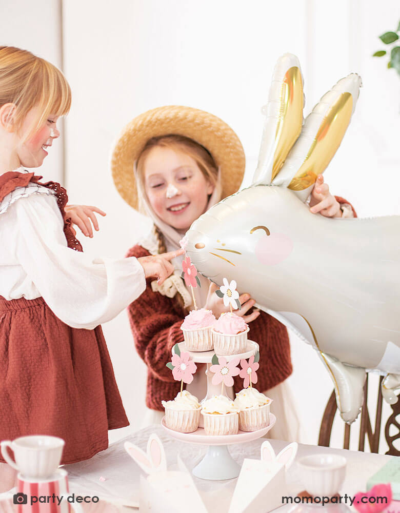 Two girls celebrating Easter with a table filled with cupcakes and treats decorated with Easter themed party supplies. The girls were also playing with Momopartys's 33x32" bunny shaped foil balloon by Party Deco with beautiful gold foil accent which sets a perfect scene for a Easter party celebration with kids.