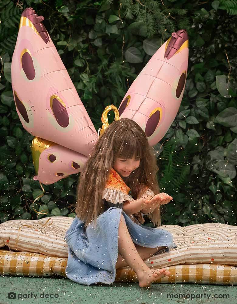 A girl sitting in a forest-like place with Momopartys's 47x 34" pastel pink butterfly foil balloon by Party Deco, with gold confetti on her hand looks like ferry's dust, it creates a whimsical vibe and look.