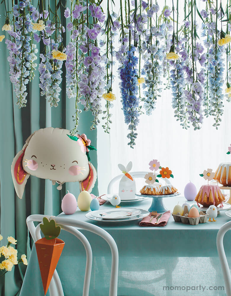 A stunning table setup for an Easter celebration featuring Party Deco's 26" floral bunny foil balloon in front of beautiful floral arrangement hanging from above; on the table it's decorated with bunny themed napkins, carrot shaped goodie boxes and bunny shaped goodie bags and flower shaped cupcake toppers