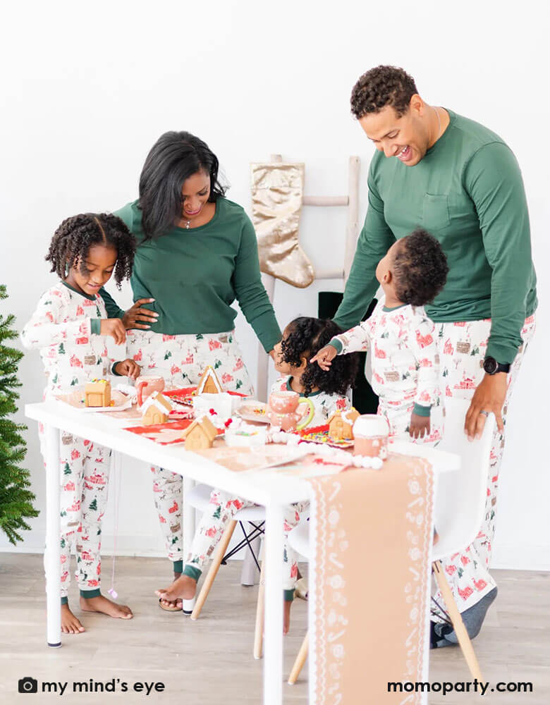 A family of 5 in their Holiday pajamas gathering at a table for a gingerbread house decorating activity featuring Momopartys's gingerbread house paper table runner draping over the table, on the tabletop is adorned with gingerbread house themed tableware including Momopartys's gingerbread house shaped plates, party napkins, gingerbread man mugs. Next to the table are some festive velvet stockings and a Christmas tree, ready for a fun kid's Holiday party!