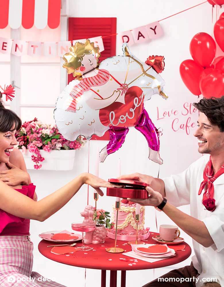 A loving couple at a dinner table enjoying a box of chocolate and pink cake to celebrate Valentine's Day. In the back the room is decorated with Momopartys's 32x34" Cupid shaped foil balloon and a bouquet of red heart shaped latex balloons, with a banner says Happy Valentine's Day in pink, it sets a romantic scene for a special Valentine's Day celebration.