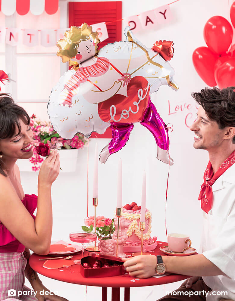 A loving couple at a dinner table enjoying a box of chocolate and pink cake to celebrate Valentine's Day. In the back the room is decorated with Momopartys's 32x34" Cupid shaped foil balloon and a bouquet of red heart shaped latex balloons, with a banner says Happy Valentine's Day in pink, it sets a romantic scene for a special Valentine's Day celebration.
