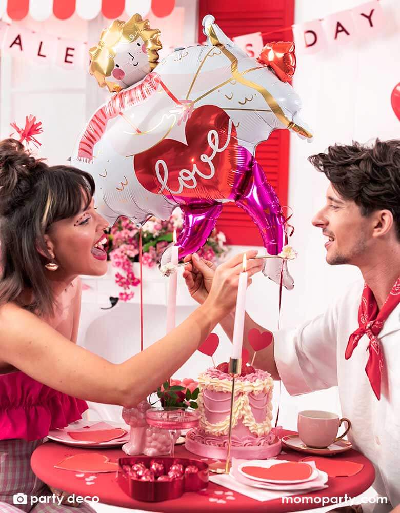 A loving couple at a dinner table enjoying a box of chocolate and pink cake to celebrate Valentine's Day. In the back the room is decorated with Momopartys's 32x34" Cupid shaped foil balloon and a bouquet of red heart shaped latex balloons, with a banner says Happy Valentine's Day in pink, it sets a romantic scene for a special Valentine's Day celebration.