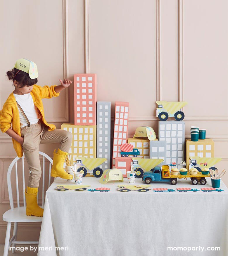 a girl wearing a yellow Construction Party Hat, a yellow sweater, she standing on a chair lifting her yellow rainboot in a Kid Dig In Construction party table. There are Meri Meri Dumper Truck Plates, Construction Party cups and Construction truck napkins, and paper made pastel color building as backdrop. what a Fun modern partware for a kid's construction themed birthday party, Dig in birthday party, big construction vehicle lover, boy's birthday party, kid's birthday party
