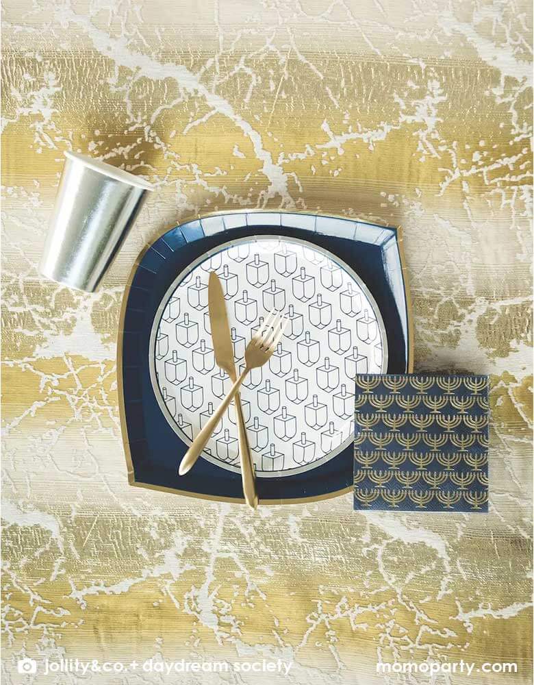 A modern and elegant Hanukkah tablescape featuring a silver party cup, a large navy napkins with gold menorah and Momopartys's 9" round plate by Jollity & Co. featuring silver foil rim and navy printed Dreidels.