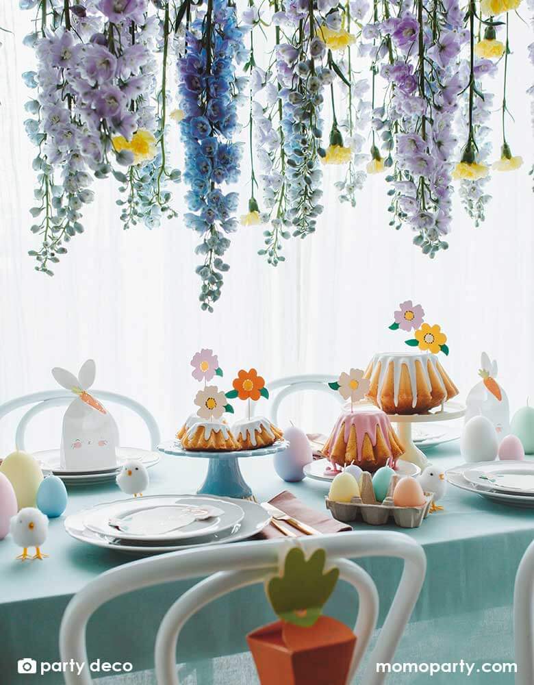 A beautiful table decorated with Easter themed tableware and decorations including Momopartys's 2.95 x 3.5 x 8.8" bunny treat bags by Party Deco, Easter chicks decorations, Easter egg shaped candles in pastel colors, and floral cupcake toppers on some bundt cakes. Above the Easter table there's wisteria flowers draping from above, makes a beautiful scene for an Easter celebration.