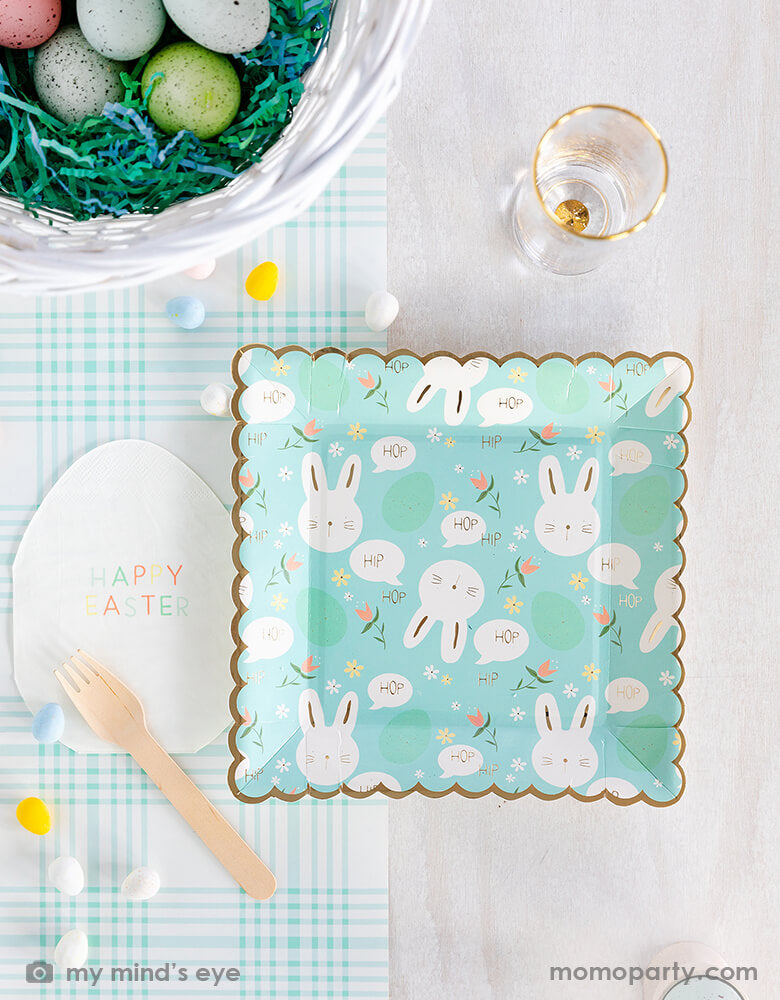 Top view of Easter party table top covered with My mind's eye - Easter Egg Shaped Napkins, Scattered Bunny Scallop Paper Plates and a wooden fork, all on a blue checkered paper table runner, surrounded by mini egg decorations and colored eggs in a green basket with mint shredded paper filling like a bird's nest a glass of drink. These fresh partyware with modern fun designs are perfect for Easter celebrations