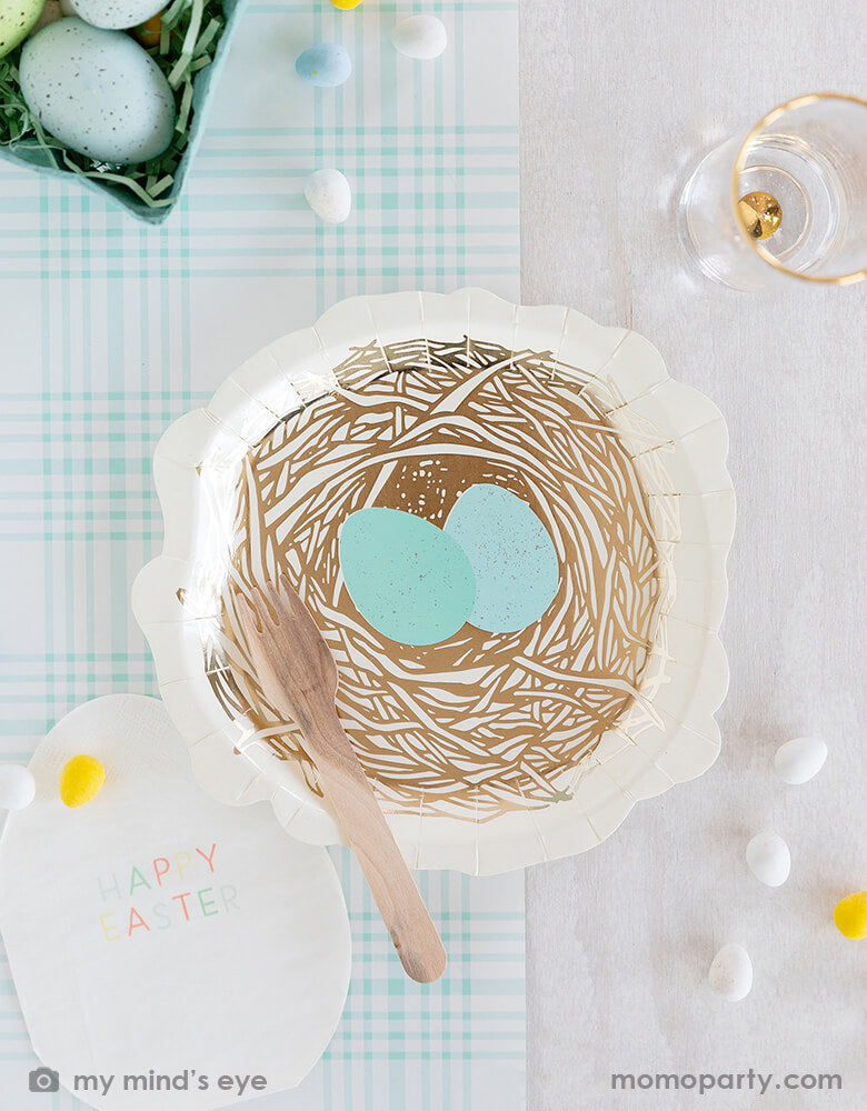 Top view of Easter party table top covered with My mind's eye -  Easter Egg Shaped Napkins, Nest shaped paper plates and a wooden fork, all on a blue checkered paper table runner, surrounded by mini egg decorations and colored eggs in a green basket with mint shredded paper filling like a bird's nest. These fresh partyware with modern fun designs are perfect for Easter celebrations