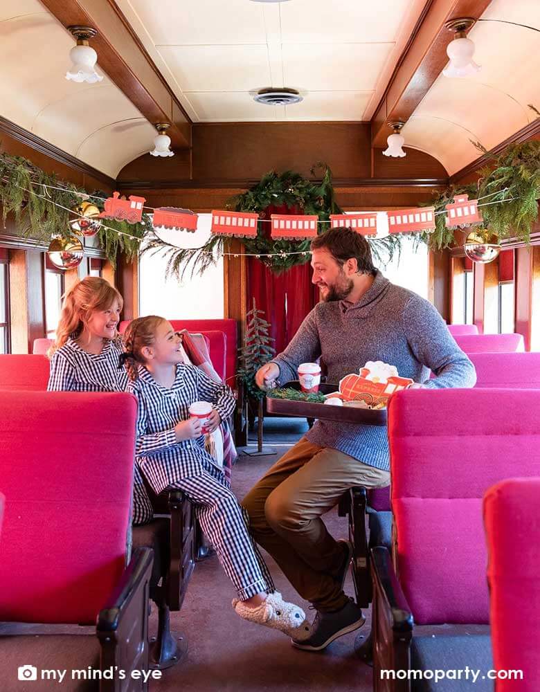 A family of a dad and two little girls in their Holiday pajamas enjoying a vintage tin tray filled with holly branches, holiday treats and cookies and Momopartys's North Pole Express train shaped plates, bell shaped side plates and polar express red 8 oz to-go cups by My Mind's Eye sitting on a vintage Polar Express train cabin decorated with Momopartys's North Pole Express train and bell garland set, a perfect idea for this Holiday season's celebration with family and kids.