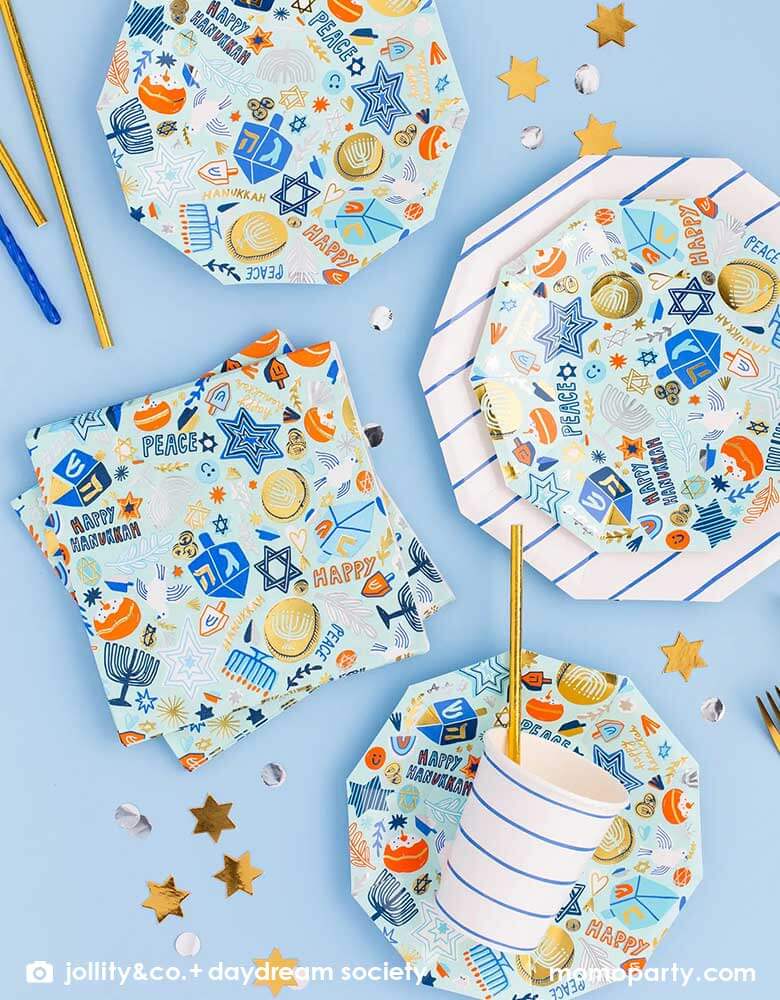 A tableware arrangement of Momopartys's 6.5" Festival of Lights Large Napkins and side plates by Daydream Society featuring Hanukkah themed symbol including dreidels, star of David, nine-branched candelabra, Menorah candles and Sufganiyah in blue colors with gold and silver foil detail, along with blue striped party cups and large plates, all together makes a modern Hanukkah tablescape